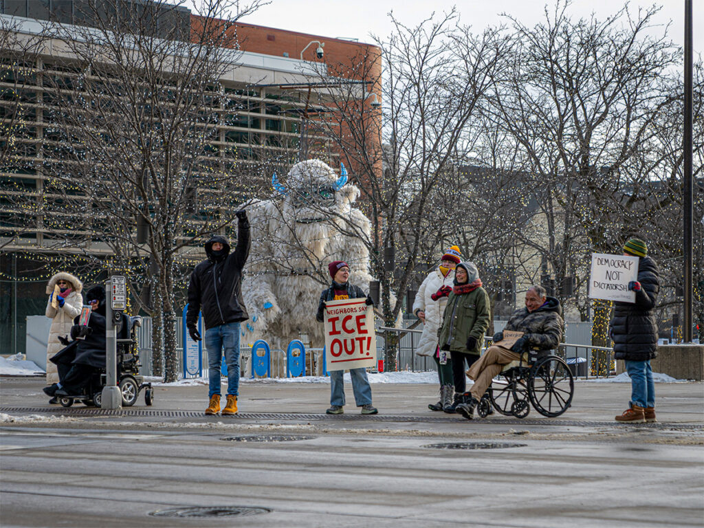 Protesters standing in the snow in front of the Holidazzle Yeti statue at South 11th Street and Nicollet in downtown Minneapolis, Minnesota, holding signs protesting ICE violence and state-sponsored killings during January 2026. (Photo by Awe Video & Photo Studio)