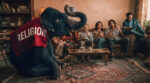 A large elephant wearing a red blanket with the word "RELIGION" printed in bold white letters sits in a living room. A diverse group of people sits on a couch behind it, laughing and distracted by their phones and books, completely ignoring the elephant’s presence. The room is cluttered with books and antique furniture, symbolizing the weight of institutional dogma.